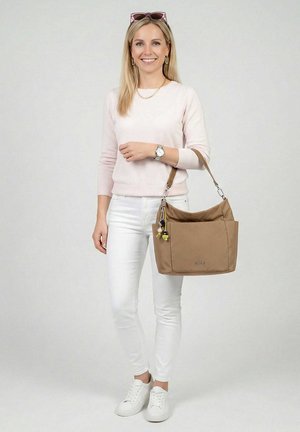 Femme debout souriante, vêtue d'un pull rose pâle et d'un pantalon blanc, tenant un grand sac à bandoulière beige avec des accessoires porte-clés.