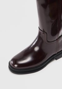 Μπορντό γυαλιστερή καουτσούκ μπότα με στρογγυλή μύτη, ανθεκτική μαύρη σόλα και διακριτική ραφή κατά μήκος του κορμού.