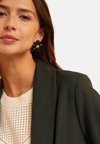 Blazer verde a quadretti con vestibilità strutturata, abbinato a un top in maglia crema con motivo ad occhielli. Orecchini neri floreali con dettagli in oro.