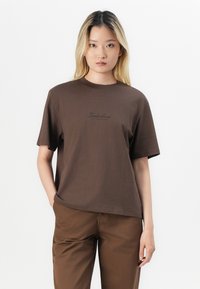 Tricou din bumbac maro, cu mâneci scurte și gât rotund, având un logo Timberland imprimat de culoare maro închis pe partea din față.