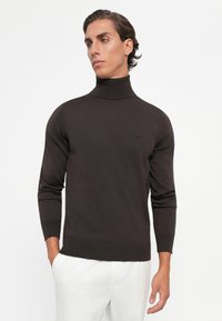Young man with dark brown turtleneck sweater and white pants, standing with one hand in pocket, looking to his left.