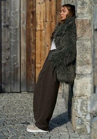 Kobieta oparta o kamienny mur, ubrana w ciemny futrzany płaszcz, szerokie brązowe spodnie, beżowe buty, na tle drewnianym i kamiennym.