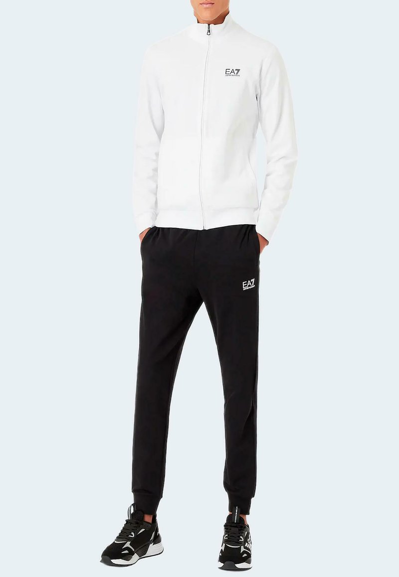 Chaqueta blanca con cremallera y cuello alto, logotipo negro; combinada con pantalones jogger negros con cinturilla elástica y logotipo a juego; ambos en tejido suave.