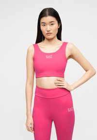 Νεαρή γυναίκα που φοράει έντονο ροζ αθλητικό σουτιέν και κολάν EA7 Emporio Armani, στέκεται με το ένα χέρι στη μέση μπροστά σε απλό φόντο.