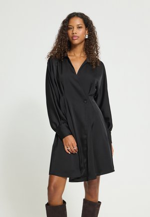 Femme aux cheveux bouclés portant une robe noire à manches longues boutonnée et des bottes foncées, debout devant un fond clair uni.