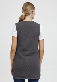Grijze gebreide vest met een gestructureerde afwerking, mouwloos ontwerp en een langere achterkant, gecombineerd met een wit T-shirt en donkere jeans.