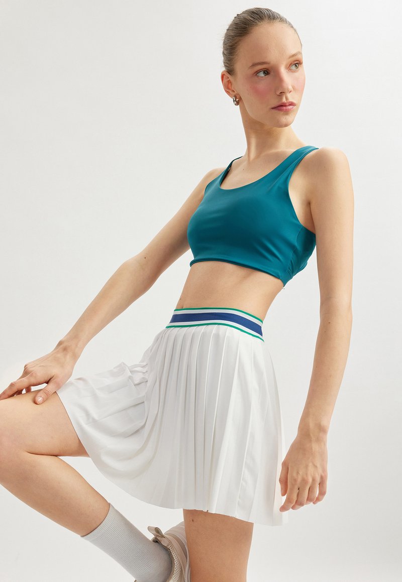 Top corto en verde azulado con un escote redondo, combinado con una falda blanca plisada que presenta una cinturilla de rayas multicolores. Textura suave, diseño atlético.