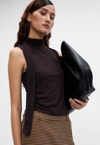 Schwarzes ärmelloses Oberteil mit hohem Kragen, kombiniert mit braunen Hahnentritt-Musterhosen. Das Model hält eine große schwarze Lederhandtasche.
