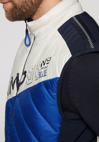 Gilet imbottito blu e bianco con alto colletto e chiusura con zip. Presenta un motivo trapuntato, tessuto in contrasto blu scuro e dettaglio del logo.