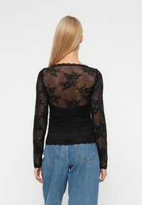 Zwarte kanten top met lange mouwen, voorzien van een doorschijnend bloemenpatroon, gather-effect bij de taille en een geschulpte halslijn. Draagt u met lichtblauwe jeans.