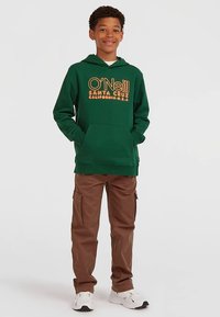 Zaļš flīsa džemperis ar kapuci un priekšējo kabatu, apdrukāts ar oranžu logotipa tekstu, komplektā ar brūnām cargo biksēm ar sānu kabatām un baltām sporta kurpēm.