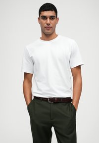 Λευκό βαμβακερό T-shirt με κοντά μανίκια, συνδυασμένο με σκούρες πράσινες παντελόνες και καφέ πλεχτή δερμάτινη ζώνη. Το μοντέλο στέκεται με τα χέρια στις τσέπες.