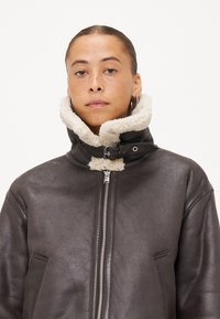 Giacca in pelle marrone scuro con colletto in shearling sintetico, chiusura con zip e tasche laterali. Texture liscia e design strutturato.