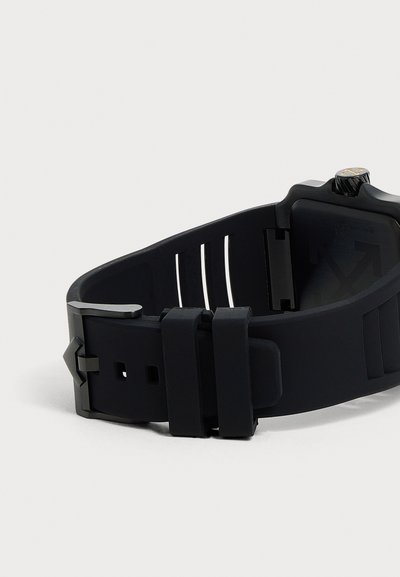 Bracelet de montre en silicone noir avec boucle métallique et trous réglables, partiellement attaché, sur un fond clair uni.