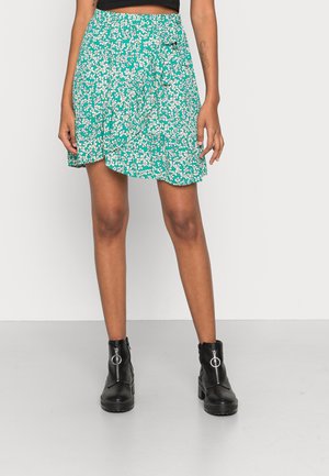 Mujer con falda cruzada verde con estampado floral por encima de las rodillas, combinada con botines negros con detalle de cremallera en forma de anillo.