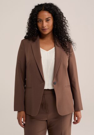 Femme aux cheveux noirs bouclés portant un blazer marron et un pantalon avec une blouse blanche, debout devant un fond beige uni.