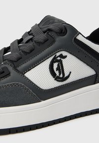 Czarno-biały sneaker z szarymi sznurowadłami, siatkową boczną częścią, skórzanym wykończeniem oraz stylizowaną czarną haftowaną literą "G" z boku.