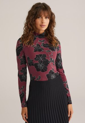 Langarm bloes met bloemenprint in bordeauxrood met zwarte bloempatronen, voorzien van een hoge hals. Gecombineerd met een geplooide zwarte rok.