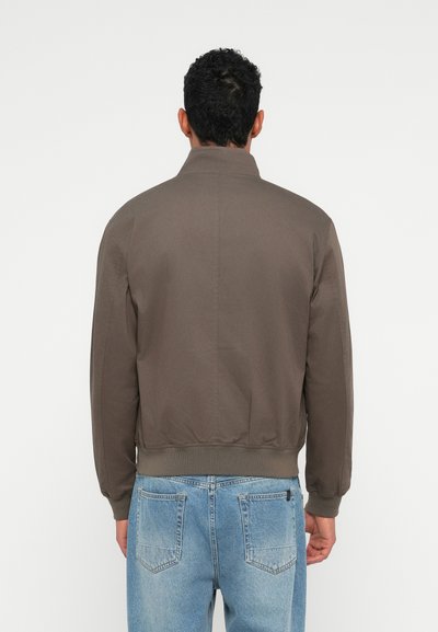 Homme portant une veste marron et un jean bleu clair, de dos contre un fond blanc uni.