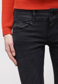 Jeans de mezclilla negra con ajuste slim, que cuentan con un bolsillo frontal, trabillas para cinturón y un suéter de punto naranja contrastante parcialmente visible.