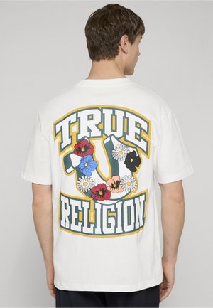 Mann trägt weißes Kurzarmhemd mit großem "TRUE RELIGION"-Schriftzug und mehrfarbigem Blumenmuster auf dem Rücken.