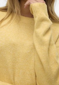 Pull jaune en tricot avec un col et des poignets côtelés. La texture est douce, avec un motif chiné subtil sur toute la surface.