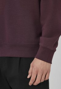 Main d'une personne portant un sweat à manches longues violet foncé et un pantalon noir, reposant à côté de leur corps contre un fond uni.