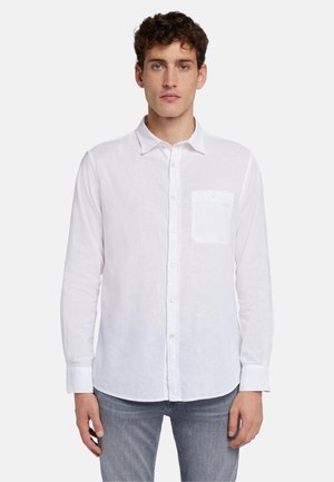 Camicia bianca in lino con taschino a sinistra, maniche lunghe e colletto, abbinata a jeans grigi chiari. Tessuto texturizzato.