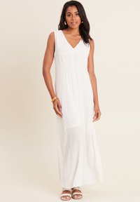 Robe maxi sans manches blanche avec un décolleté en V, tissu texturé, taille froncée et silhouette fluide. Présente des sandales plates et des bijoux en or.