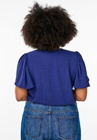 Blouse violette avec des manches à volants, présentant une texture subtile à motifs animaliers, associée à un jean en denim bleu taille haute.
