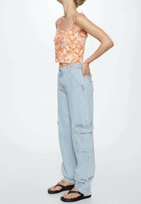 Top corto floreale in arancione e rosa con bordi arricciati; jeans cargo oversize azzurri chiari con tasche; infradito nere con fasce.