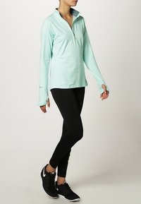 Kobieta ubrana w jasnoniebieską bluzę zapinaną na zamek z długimi rękawami z otworami na kciuki, czarne legginsy oraz czarne sneakersy Nike, stojąca na jednej nodze.