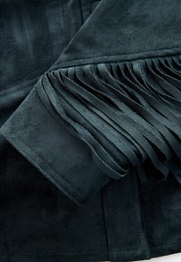 Dunkelgrüner Wildlederstoff mit geschichteten Fransen entlang des Bündchens, der eine glatte Textur und detaillierte Nähte aufweist, und ein modernes Design präsentiert.