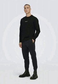 Man wearing black long-sleeve shirt with small logo, dark pants, and black boots, standing with one hand in pocket on plain background.