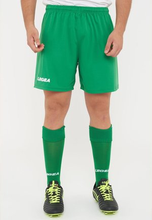 Mężczyzna w zielonych szortach sportowych, dobranych do nich wysokich skarpetach i czarnych korkach z żółtymi sznurowadłami, stojący na jednolitym tle.