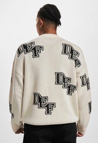 Cremfarbener Pullover mit lockerer Passform, der mit auffälligen schwarzen "DEF"-Buchstaben durchzogen ist, gerippten Bündchen und einem strukturierten Strickdesign.