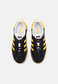 Zapatillas de ante negro con tres franjas amarillas, forro interior blanco, cordones negros y suela de goma. Incluye una etiqueta de marca negra en la lengüeta.