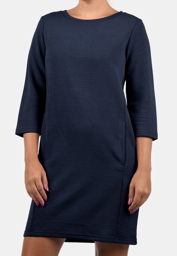 Femme portant une robe texturée bleu marine à manches longues, longueur genoux, avec un col rond, debout devant un fond uni.