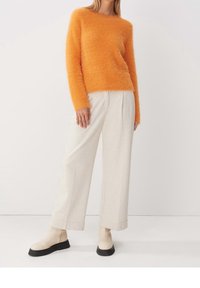Oranje pluizige sweater met lange mouwen, gecombineerd met lichtbeige wijde broek. Schoenen zijn crème met een zwarte zool. Effen achtergrond.