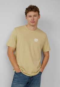Beige katoenen t-shirt met korte mouwen en een ronde hals, met een klein wit 'Evolving Roots' logo op de linkerkant van de borst.