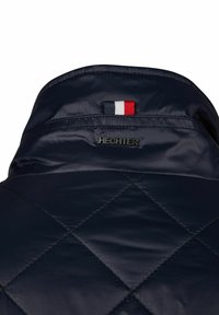 Navyblaue gesteppte Jacke mit einem Kragen, versehen mit dem HECHTER-Logo und einem kleinen schwarzen, weißen und roten Stoffetikett, das zentriert darüber angebracht ist.