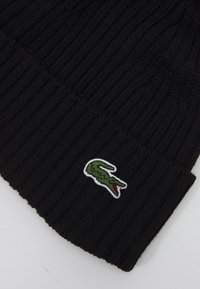 Svart stickad mössa med ribbad textur. Har en vitkantad grön krokodil-logga framtill. Enkelt och mångsidigt design.