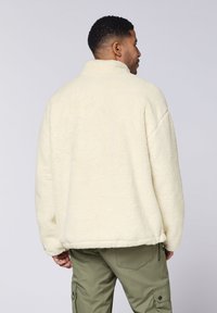 Cremfarbener Fleece-Pullover mit hohem Kragen, großzügigem Schnitt und weicher Textur, kombiniert mit olivgrünen Cargo-Hosen.