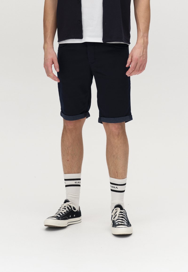 Marineblaue Shorts mit umgeschlagenen Säumen, kombiniert mit schwarzen Sneakers und weißen Socken mit schwarzen Streifen. Lässiger, leichter Stoff.