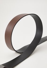 Curea din piele texturată maro și neagră, cu o finisare netedă și un design plat, expusă într-o formă artistică pe un fundal alb.