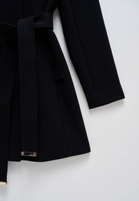 Manteau noir à manches longues avec une ceinture en tissu nouée à la taille, posé à plat sur une surface blanche.