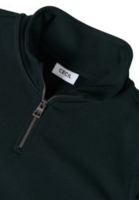 Pull à col zippé quart vert foncé avec fermeture éclair argentée et étiquette intérieure blanche indiquant "CECIL" sur le col haut.