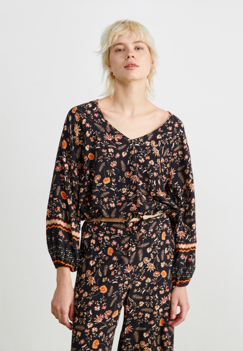 Blouse florale noire à manches longues, col en V, présentant des motifs orange, rouge et vert, avec des poignets froncés et une coupe décontractée. Ceinturée à la taille.