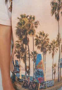 Koszulka z krótkim rękawem z grafiką palm i graffiti, wykonana z miękkiego materiału na pastelowo-różowym tle.