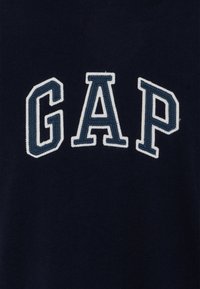 Námořnická modrá bavlněná mikina se bílým vyšitým nápisem "GAP" v tučném písmu. Text má mírně vyvýšenou strukturu.